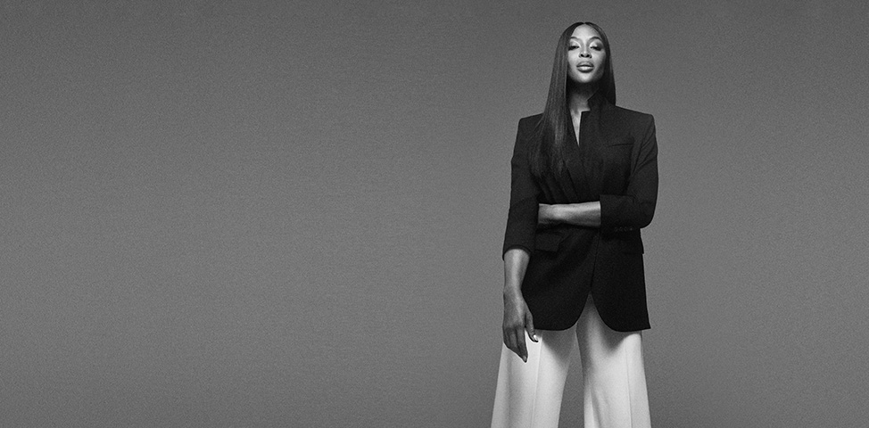 Black and white foto of Naomi Campbell with a black suit jacket and white wide pants (Photo)